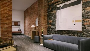 A modern room with a brick wall, a grey sofa, a small table with a lamp, a suitcase, and a framed picture on the wall. - Nester Finn Square Victoria (Montréal)