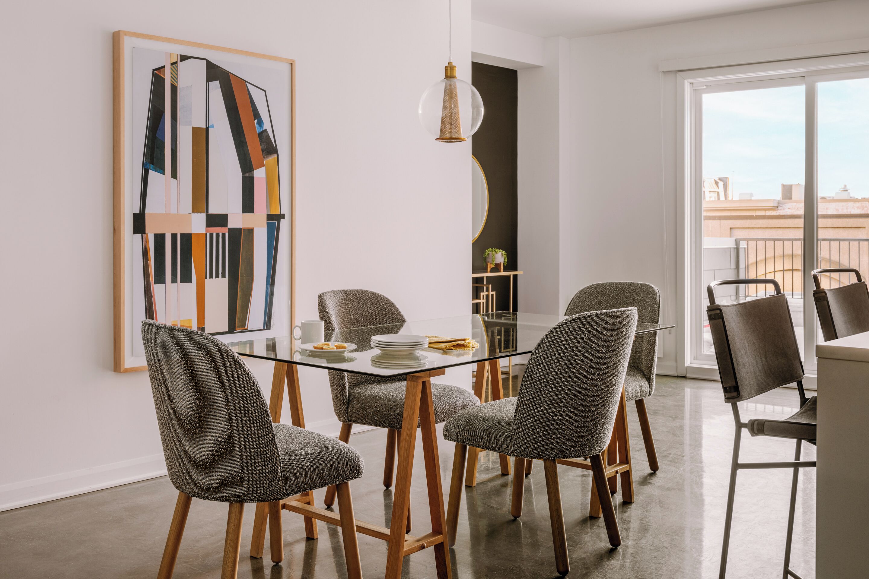 A modern dining area with a glass table, grey upholstered chairs, and a large abstract painting on the wall.
