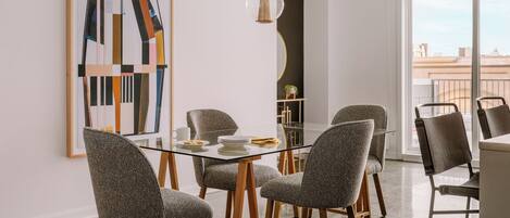 A modern dining area with a glass table, grey upholstered chairs, and a large abstract painting on the wall.