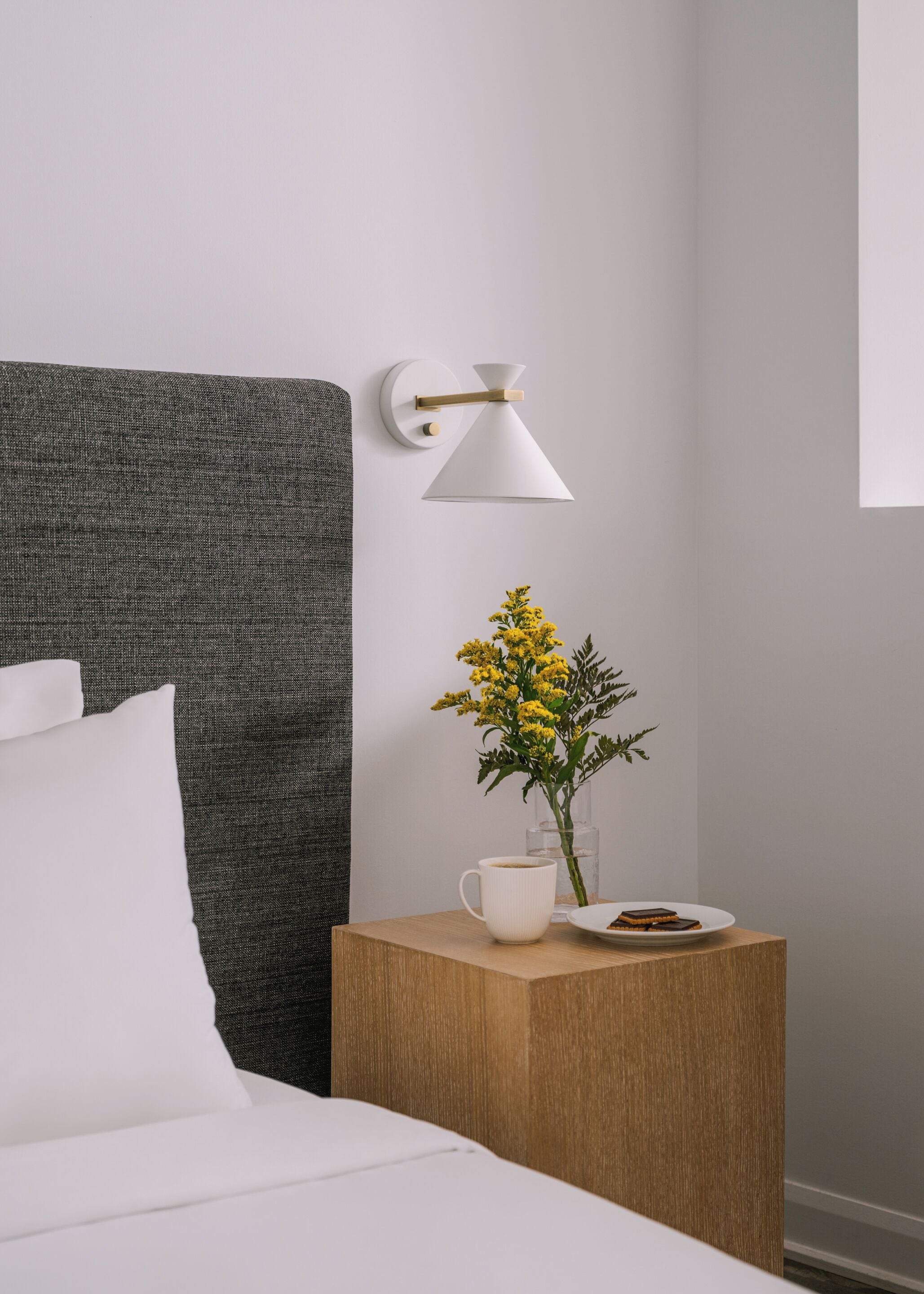 A neatly made bed with white bedding, a small wooden nightstand with a white mug, a plate with a dessert, and a vase with yellow flowers.