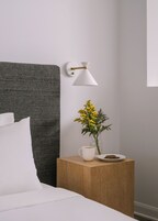 A neatly made bed with white bedding, a small wooden nightstand with a white mug, a plate with a dessert, and a vase with yellow flowers.
