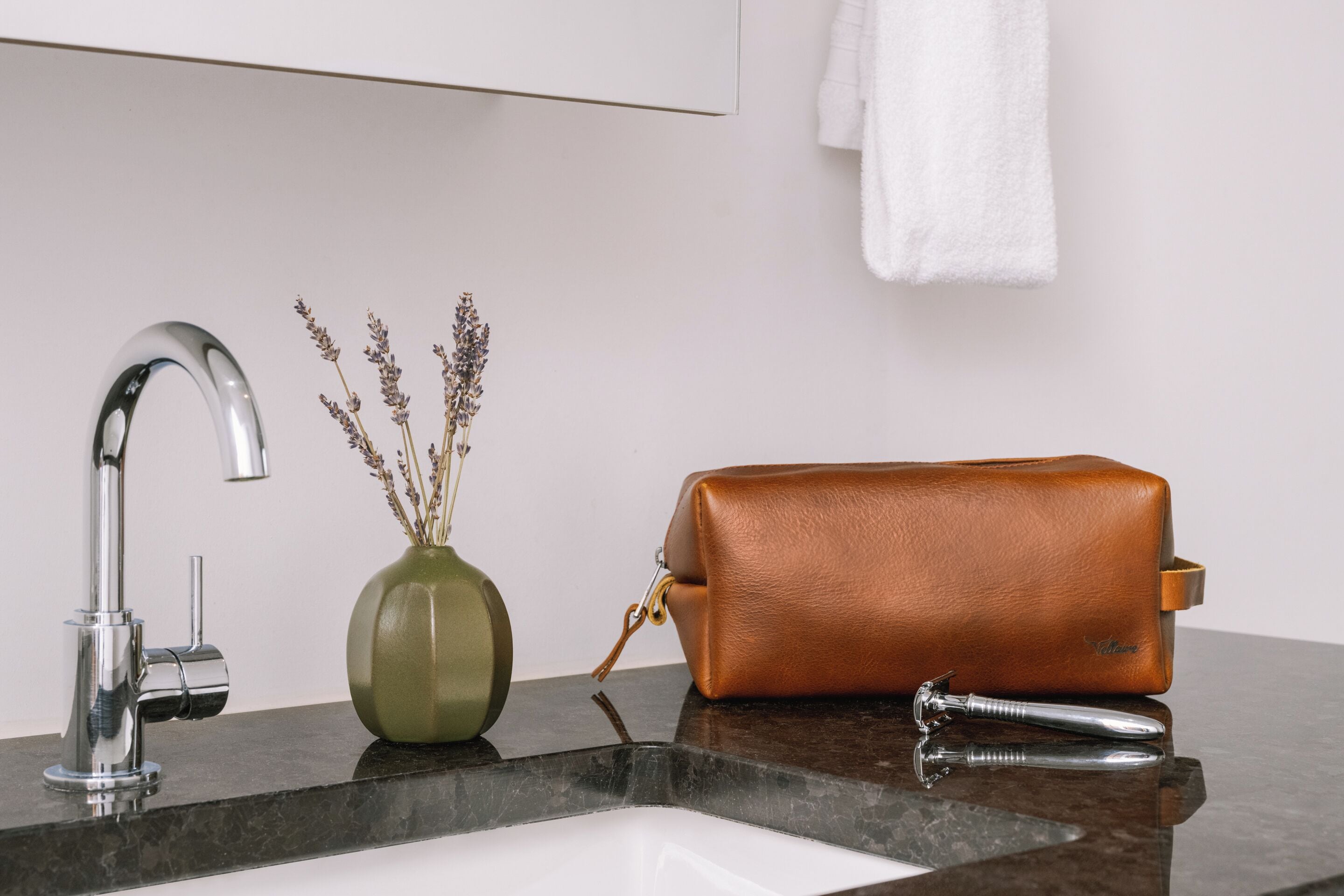 A bathroom with a modern faucet, a vase with dried flowers, and a brown leather bag on the counter.