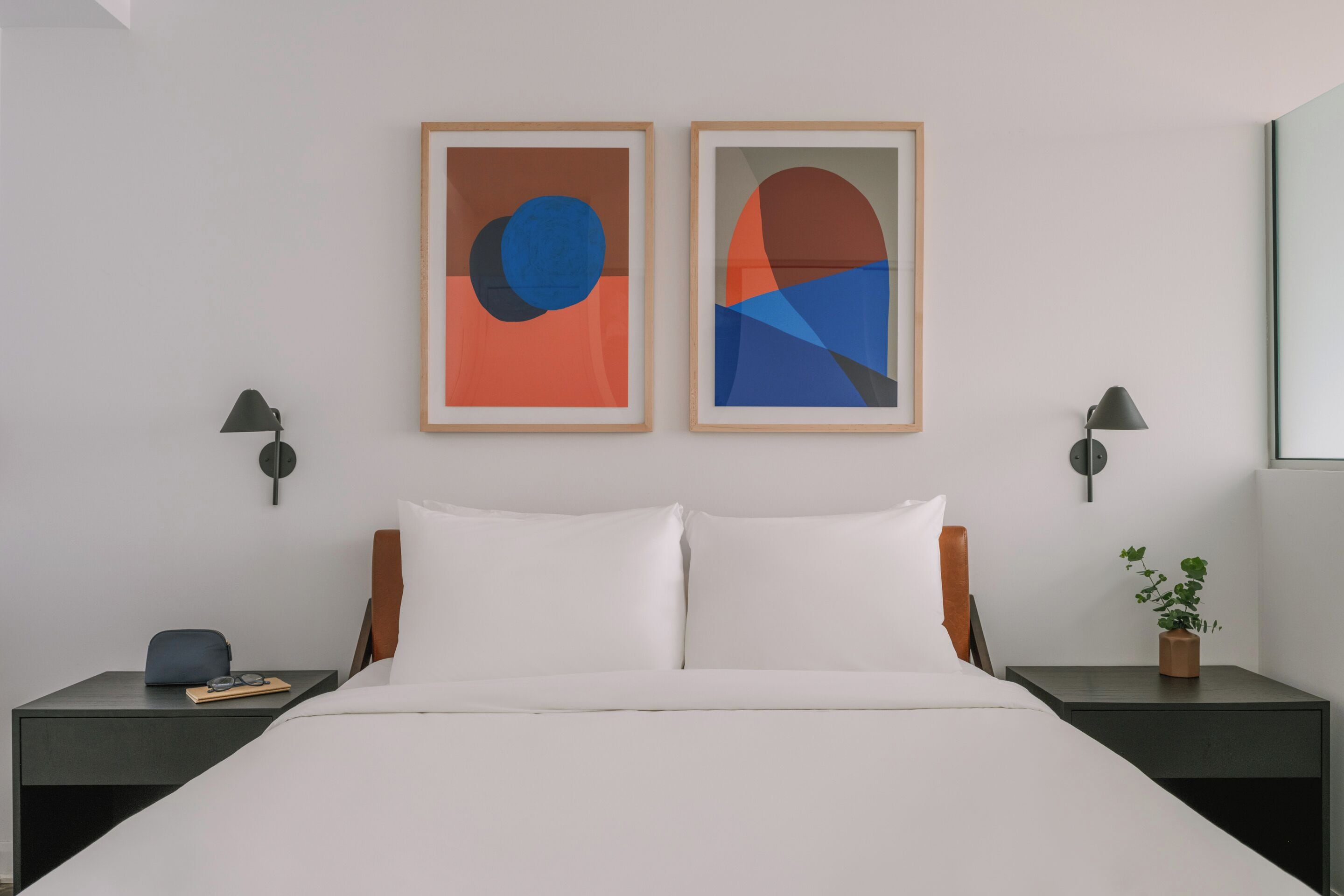 A modern hotel room with a bed, two framed abstract paintings, bedside lamps, a nightstand with a plant, and a small table with glasses and a notebook.