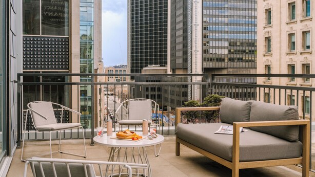 A balcony with a sofa, chairs, and a small table with fruits and drinks. - Nester Finn Square Victoria (Montréal)