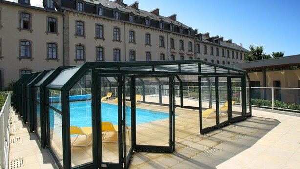A covered outdoor swimming pool with glass walls, a clear blue pool, and yellow lounge chairs. - Dinan Stay With Free Parking (Dinan)
