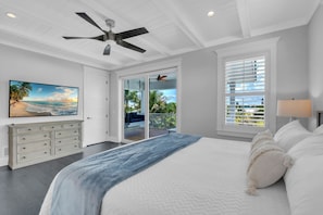 A bedroom with a bed, a ceiling fan, a large window with blinds, a painting, and a sliding door leading to a balcony. - Slice of Paradise by Duncan Real Estate (Holmes Beach)