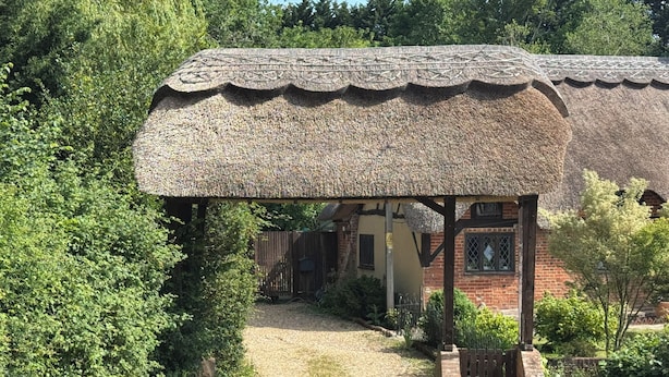 Ein Gebäude mit einem Reetdach und einer Holzveranda, umgeben von viel Grün, mit einem Backsteinhaus im Hintergrund.