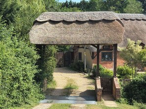 Ein Gebäude mit einem Reetdach und einer Holzveranda, umgeben von viel Grün, mit einem Backsteinhaus im Hintergrund.