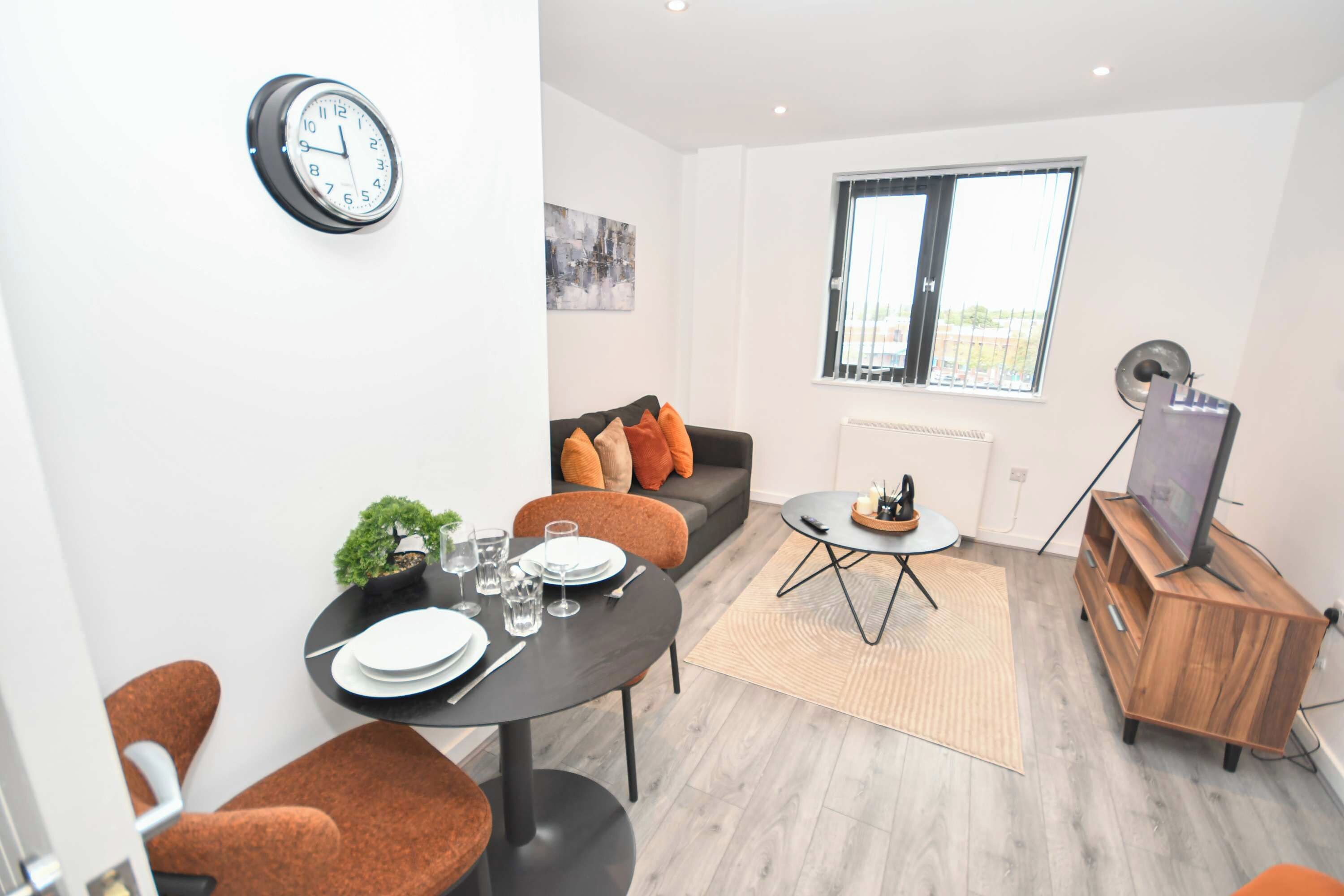 A modern living room with a dining table set for two, a sofa with cushions, a small side table, a television on a wooden stand, and a clock on the wall.