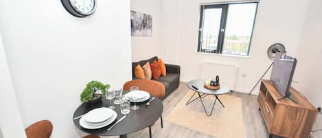 A modern living room with a dining table set for two, a sofa with cushions, a small side table, a television on a wooden stand, and a clock on the wall.