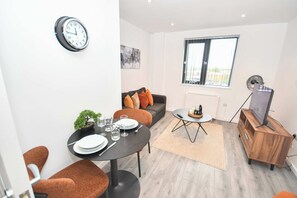 A modern living room with a dining table set for two, a sofa with cushions, a small side table, a television on a wooden stand, and a clock on the wall. - Lovely 1 Bedroom Apartment in Leeds w WiFi (Leeds)
