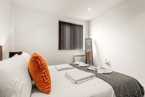 Habitación de hotel con cama, almohadas, una ventana con cortinas, una mesita de noche, un espejo y una lámpara de pared.