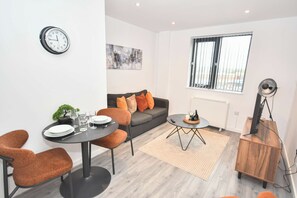 A modern living room with a dining area, a gray sofa with orange cushions, a round coffee table, and a clock on the wall. - Lovely 1 Bedroom Apartment in Leeds w WiFi (Leeds)