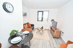 A modern living room with a dining area, a sofa with cushions, a coffee table, a TV on a wooden stand, and a wall clock. - Lovely 1 Bedroom Apartment in Leeds w WiFi (Leeds)