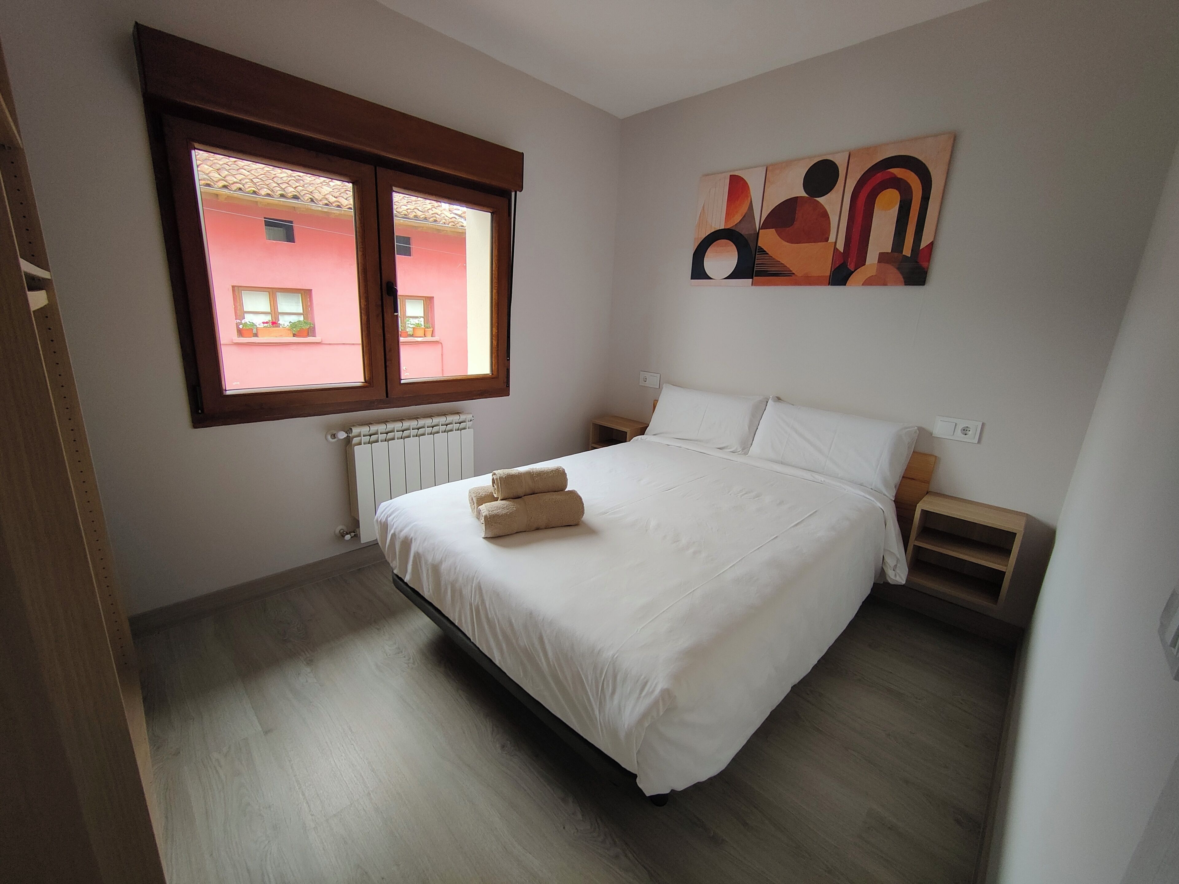Un dormitorio con una cama, una ventana, un radiador, una mesita de noche y un cuadro en la pared.