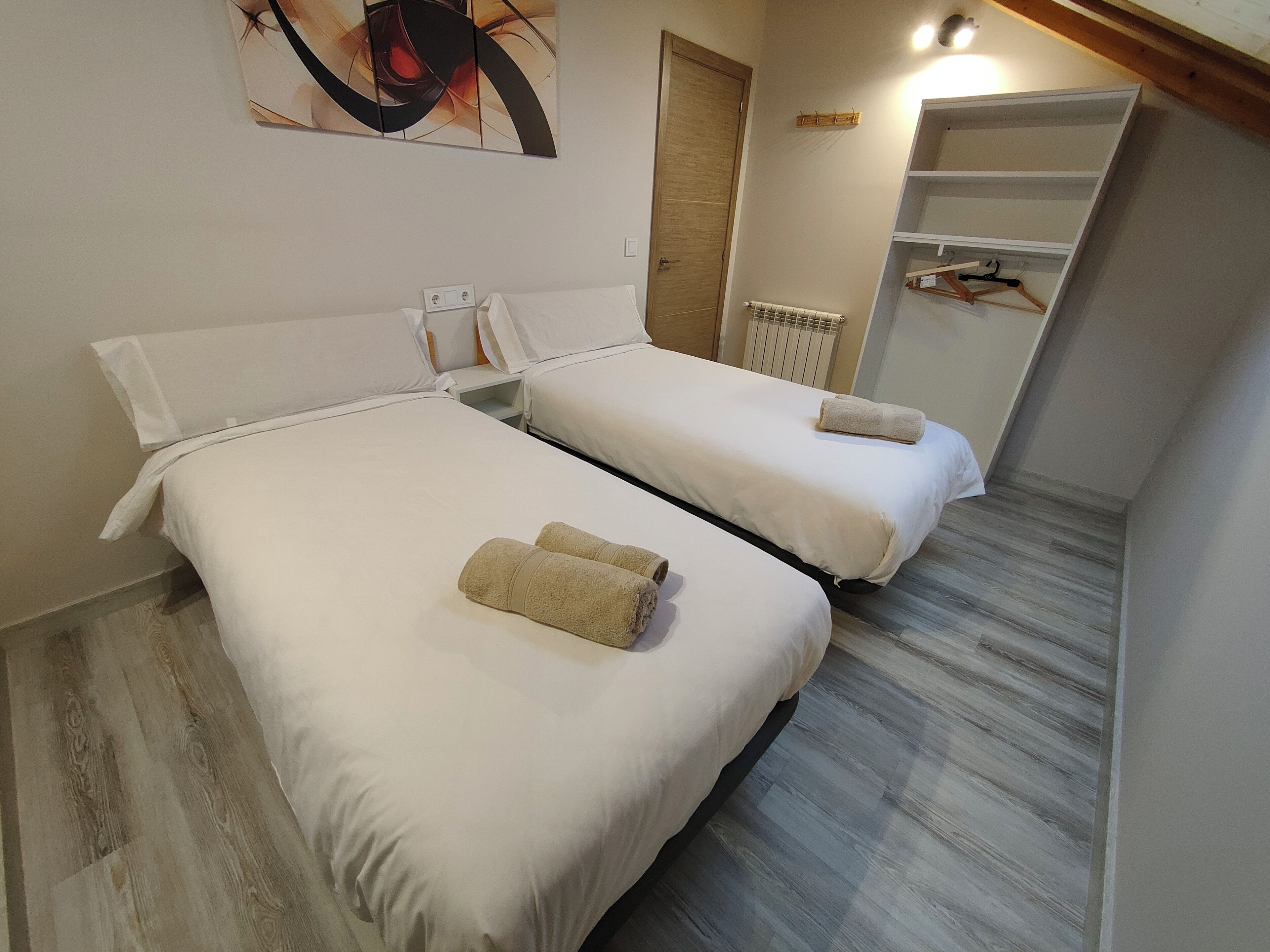 Dos camas individuales con sábanas y toallas blancas, suelo de madera, un radiador y una estantería en la pared.