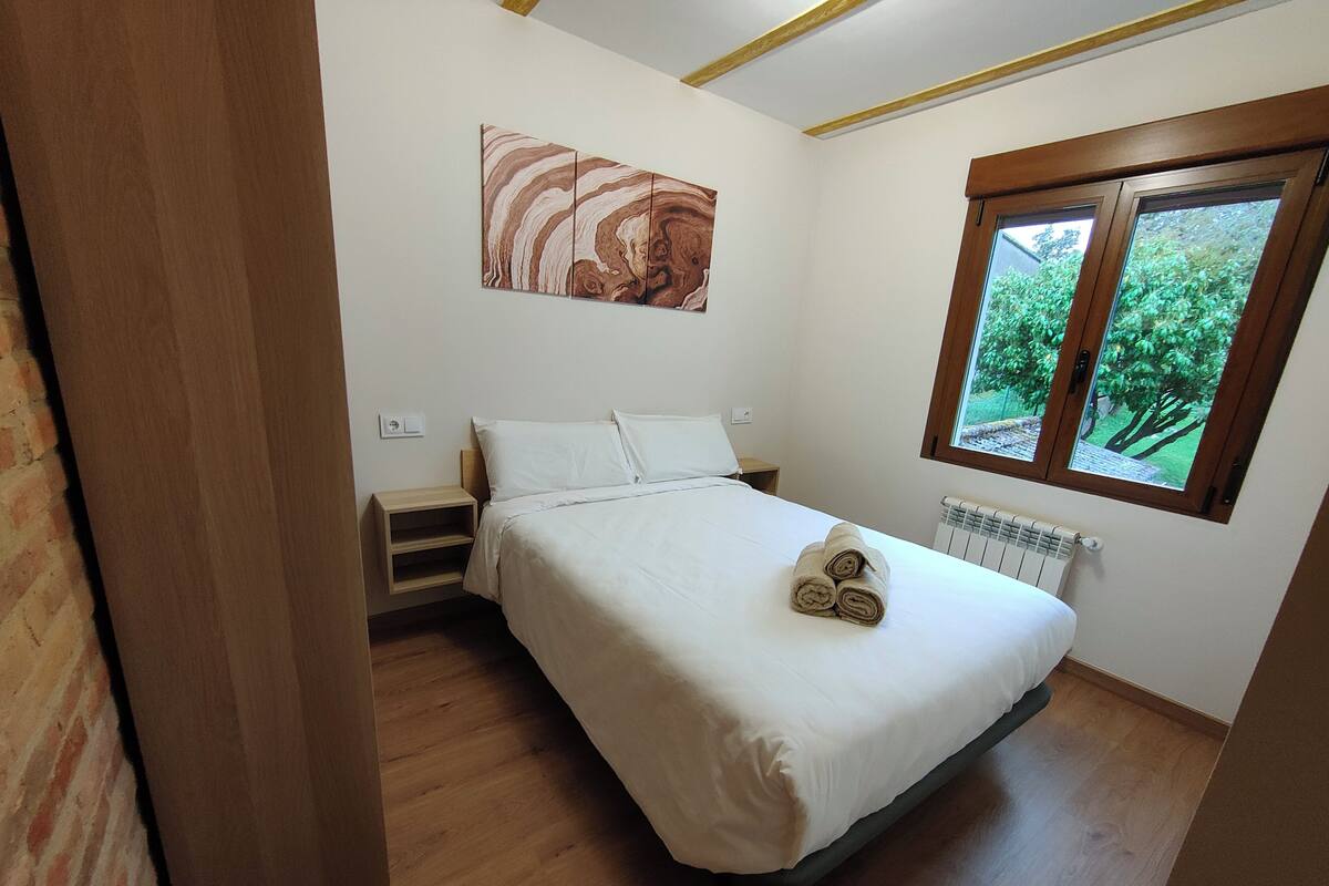 A bedroom with a bed, a nightstand, a radiator, and a window with a view of greenery.