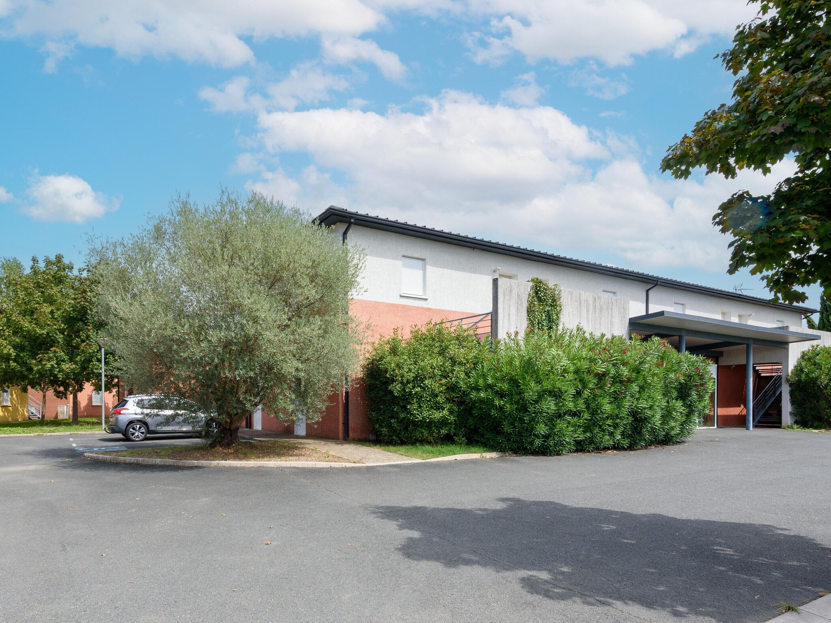 A two-story building with a garage, a covered parking area, and a tree in front.