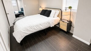 Une chambre à coucher comprenant un lit, des tables de chevet, une lampe, une fenêtre avec des stores et une porte donnant sur une autre pièce.
