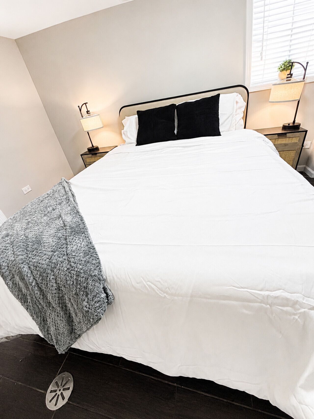 A neatly made bed with a white duvet, a grey throw blanket, and a black pillow. There are two bedside tables with lamps, a window with blinds, and a small potted plant on the right.
