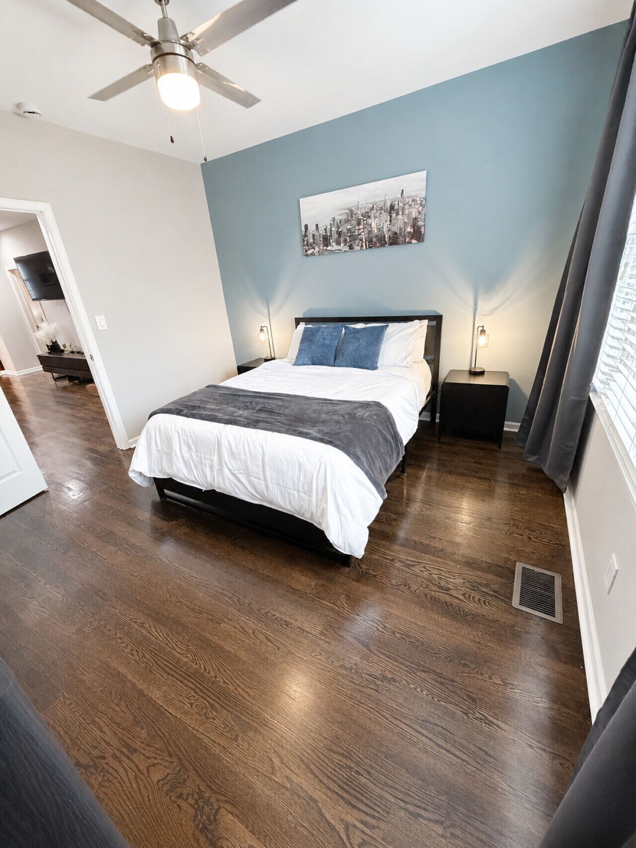 A bedroom with a bed, a ceiling fan, a nightstand, a wall-mounted light, and a window with curtains.