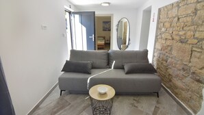 Una sala de estar moderna con un sofá gris, una mesita redonda con un tazón, una pared de piedra y una puerta que da a otra habitación.