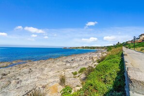 A coastal landscape with a rocky shoreline, a paved walkway, and buildings in the background. - Casa Vacanza Anika Portoscuso – Apartment with Sea View and Garden (Portoscuso)