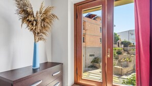 Quarto com um armário de madeira, um vaso azul com plantas secas e uma porta de vidro deslizante que dá para área externa com edifícios e vegetação.
