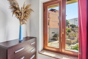 A room with a wooden cabinet, a blue vase with dried plants, and a sliding glass door leading to an outdoor area with buildings and greenery. - Casa Vacanza Anika Portoscuso – Apartment with Sea View and Garden (Portoscuso)