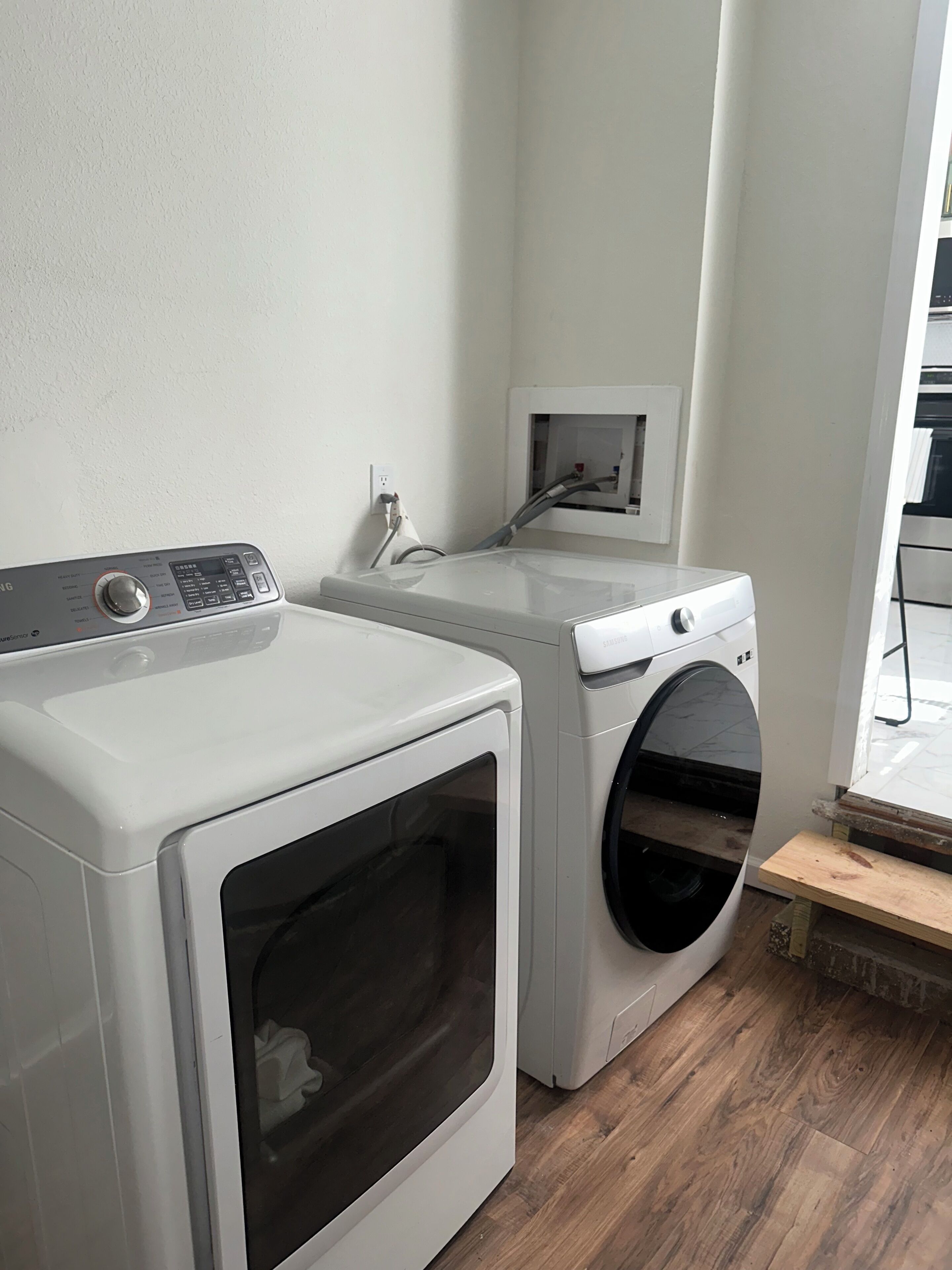 Een wasserette met twee witte wasmachines, een klein houten bankje en een stopcontact op de muur.
