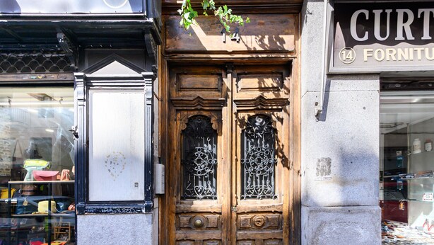 Une porte à double battant en bois, ornée de motifs métalliques travaillés, une enseigne au-dessus, et le reflet d’un intérieur coloré sur la vitre de la porte.
