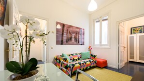 Une pièce comprenant une table à manger, un canapé recouvert d’un tissu aux motifs géométriques colorés, un tableau au mur et une plante sur la table.