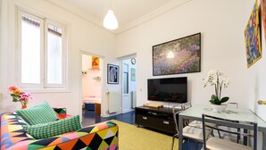 Un salon moderne avec un canapé coloré, un téléviseur à écran plat, une table à manger avec des chaises et une grande fenêtre équipée de stores.