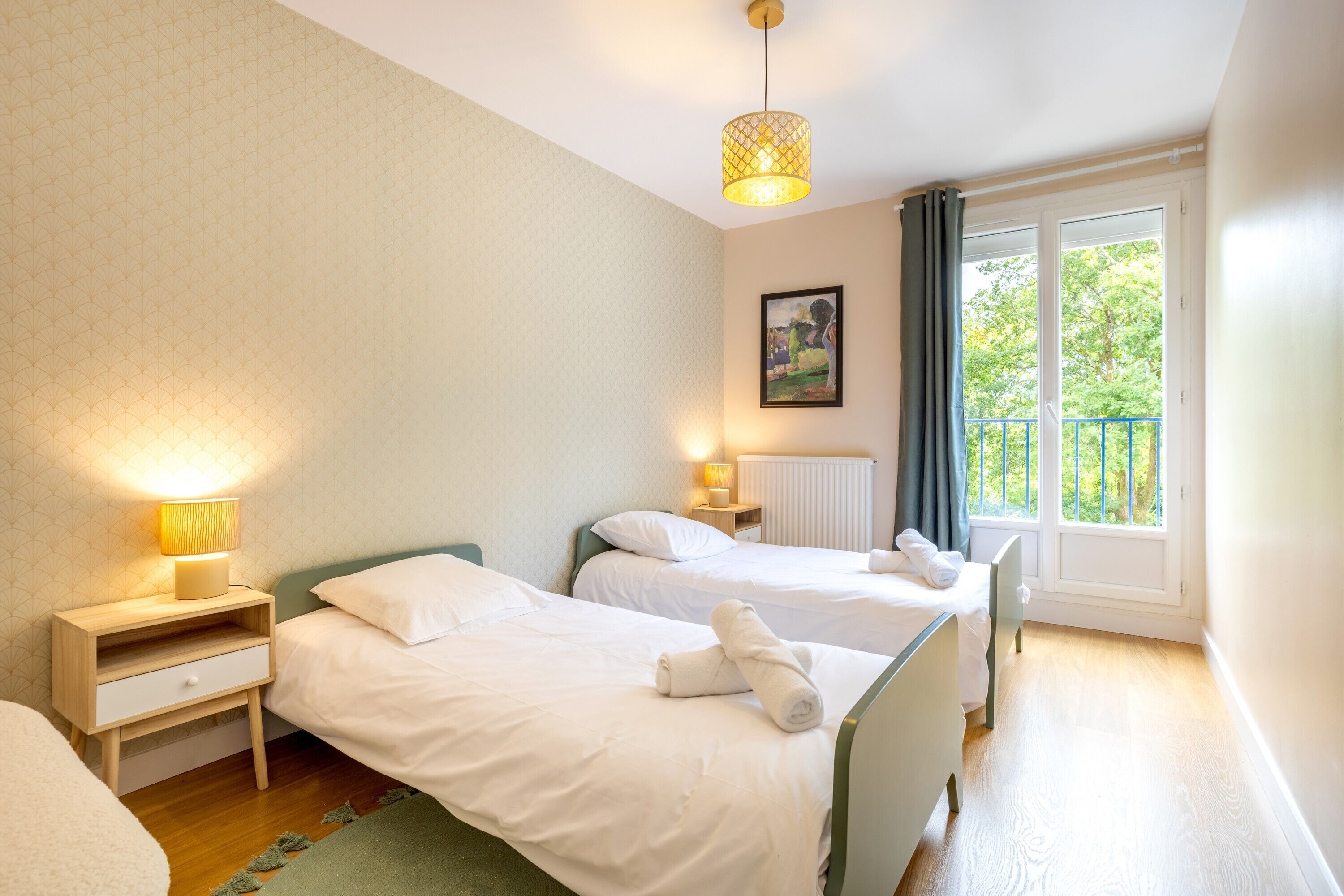 A hotel room with two beds, a nightstand with a lamp, a painting on the wall, and a window with a view of greenery.