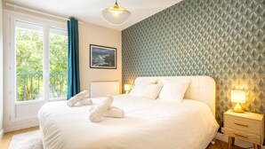 A neatly made bed with white linens and rolled towels, a bedside table with a lamp, a framed picture on the wall, and a window with curtains. - Apartment “Ty Heol – Balcony and Near Center” with Balcony and Wi-Fi (Quimper)