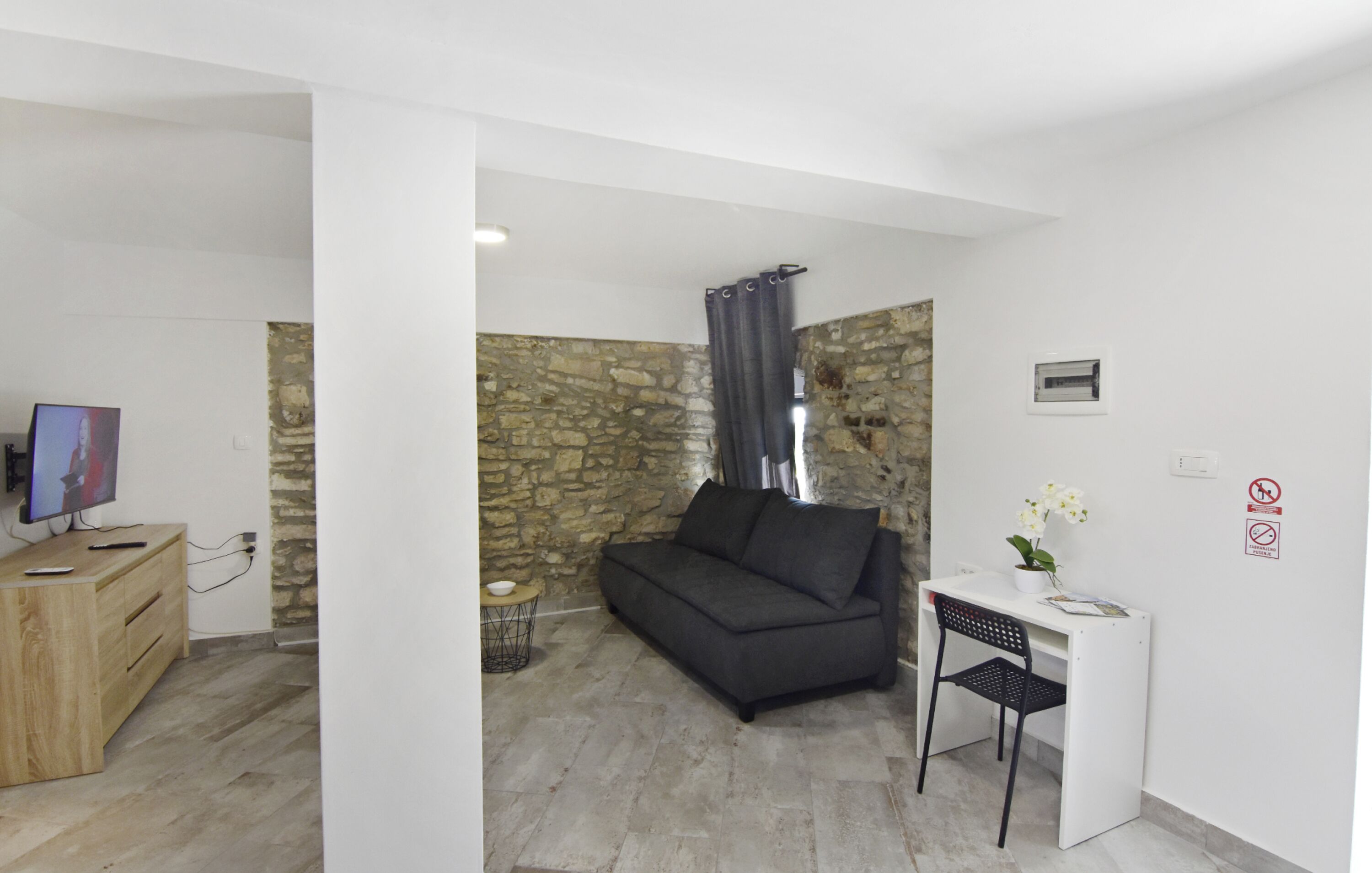 A modern living room with a stone wall, a dark sofa, a small desk with a chair, and a television on a wooden cabinet.