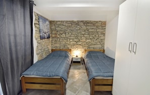 Een slaapkamer met twee houten bedden, een stenen muur, een nachtkastje, een witte inloopkast en een gordijn.
