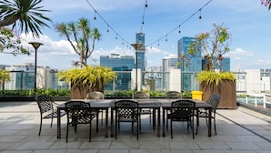 Uma área de refeições ao ar livre com vista para o horizonte da cidade, com mesa, cadeiras, plantas em vasos e luzes de cordão.
