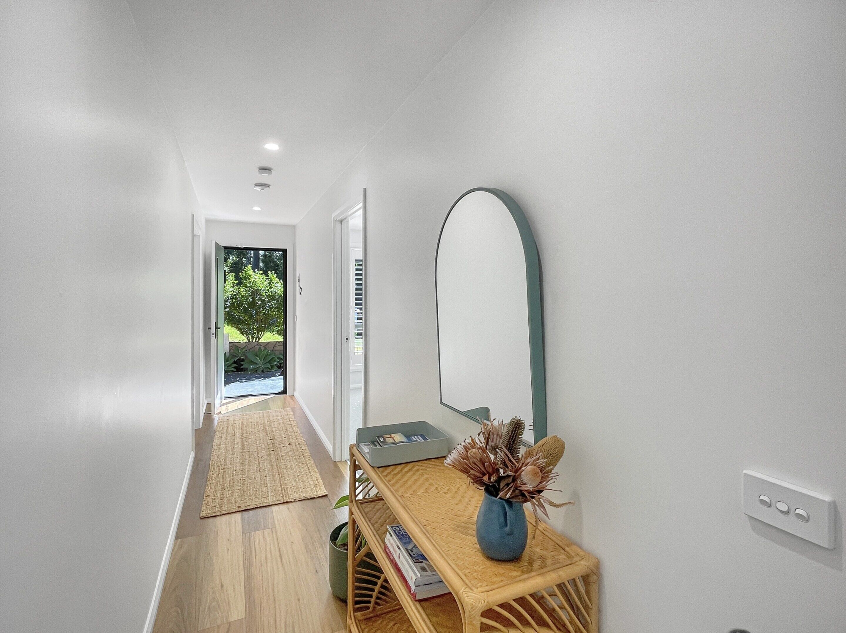 A hallway with a wooden floor, a woven rug, a small table with a vase, and a large mirror on the wall.