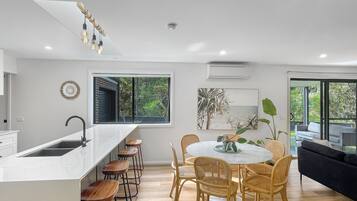 A modern kitchen with a central island, bar stools, a dining table with chairs, and a large window with a view of greenery.