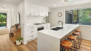Uma cozinha moderna com armários brancos, uma ilha central com banquetas, uma janela grande com vista para o verde e uma luminária pendente.