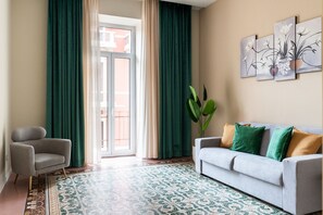 Une pièce avec un canapé gris, une plante verte, un tapis à motifs et une fenêtre avec des rideaux.