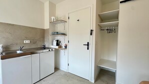 A compact kitchen with a sink, stove, and shelves; a white door with a handle; and a closet with hanging space. - StationBeds Studio Station&citycentre (Alkmaar)