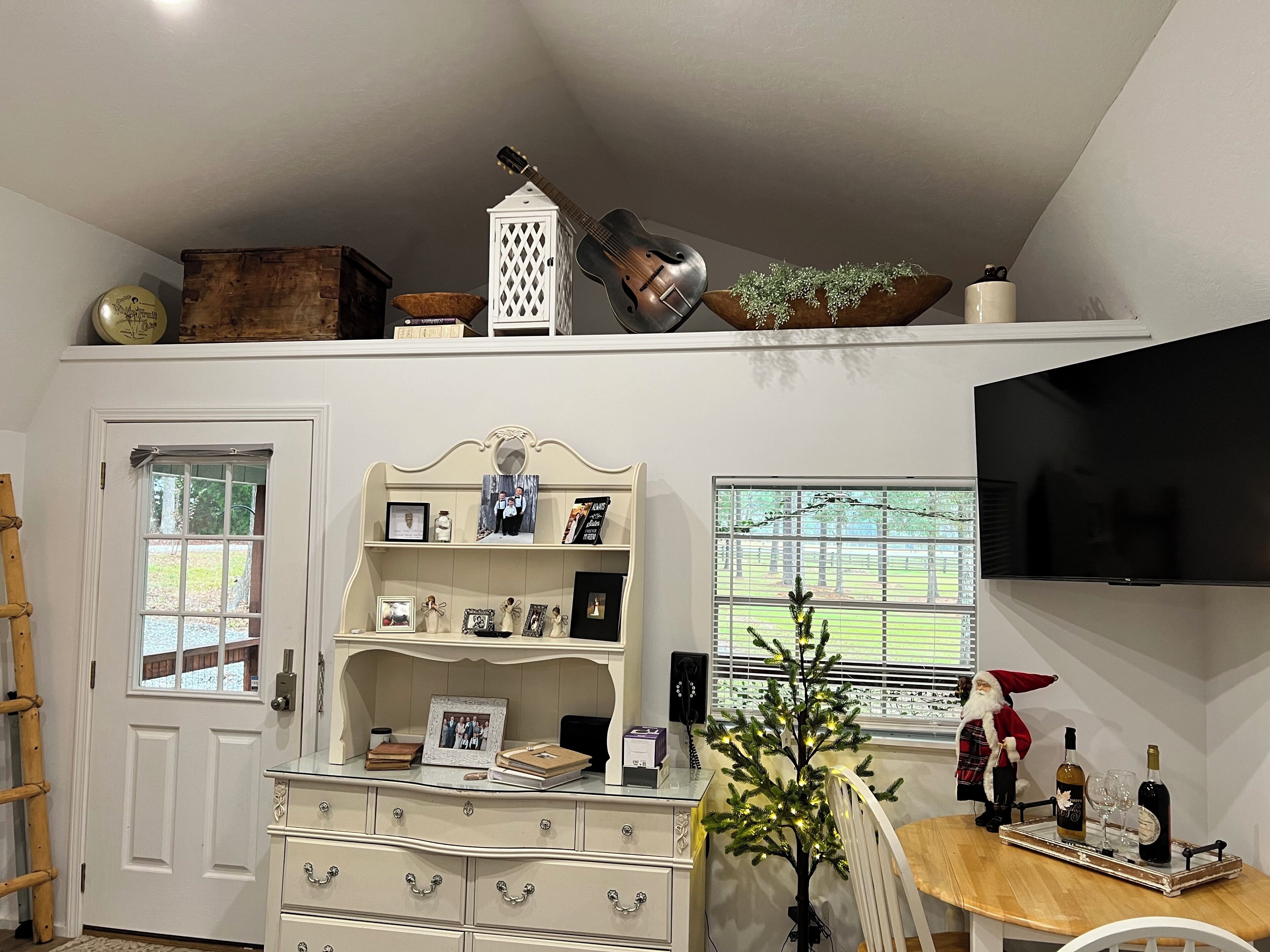 A living room with a TV, a Christmas tree, a wooden table with bottles, and a white cabinet with drawers.