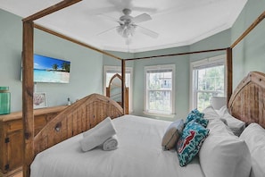 Ein Schlafzimmer mit einem hölzernen Kopfende, einem Deckenventilator, einem großen Fenster und einem Strandbild.