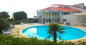 A swimming pool with lounge chairs, a palm tree, and a building with a red roof. - Le Petit Boyard – swimming pool, family and beach (Fouras)