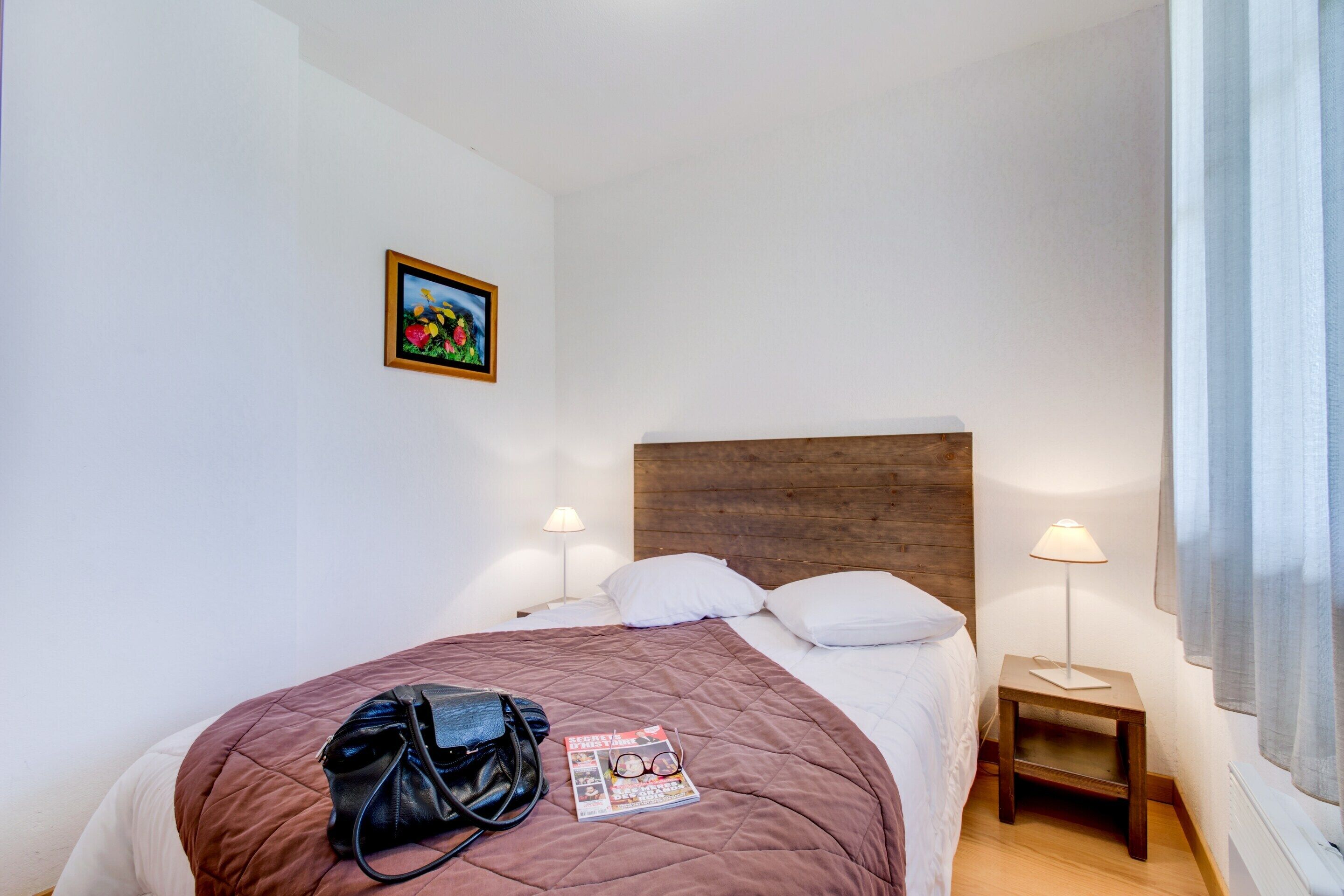 Una cama bien hecha con colcha, una revista y un bolso encima. Hay dos lámparas de noche, una mesita y un cuadro en la pared.