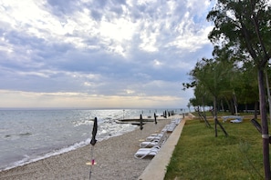 Ein Kiesstrand mit Liegestühlen und Sonnenschirmen, eine begrünte Fläche mit Bäumen und Meerblick unter bewölktem Himmel.