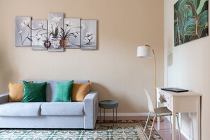 Een woonkamer met een bank, een klein rond tafeltje, een bureau met stoel en een lamp.