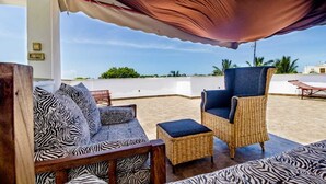 An outdoor seating area with wicker furniture and patterned cushions, a covered section, and a view of the beach. - Taraji Suites Diani (Diani Beach)